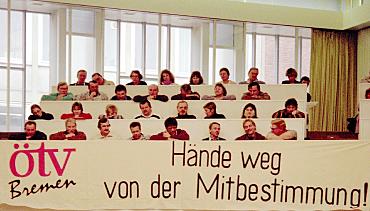 Das Bild zeigt etwa 20 Menschen, die in einem Saal  sitzen. Vor ihnen hängt ein Transparent, auf dem zu lesen ist: ÖTV Bremen - Hände weg von der Mitbestimmung.