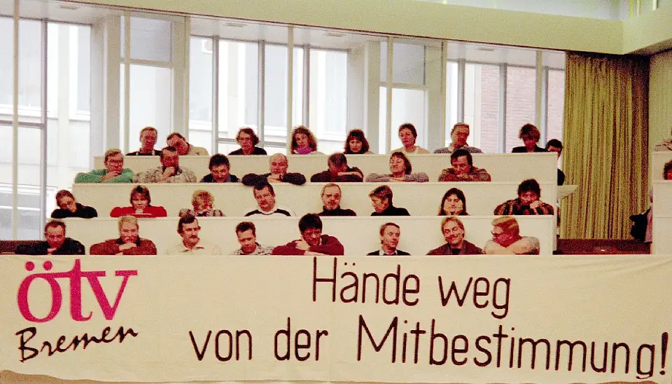 Das Bild zeigt etwa 20 Menschen, die in einem Saal  sitzen. Vor ihnen hängt ein Transparent, auf dem zu lesen ist: ÖTV Bremen - Hände weg von der Mitbestimmung.