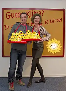 2 Beschäftigte halten ein Tablett mit vielen Smileys, die verschiedene Gesichtsausdrücke haben