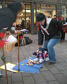 Schauspiel: Ältere Erzieherin versucht angetrengt, ein Kind (Puppe) vom Fußboden auf den Arm zu nehmen.