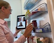 Eine Frau an einer geöffneten Kühlschranktür. In der einen Hand hält sie ein Tablet, in dem eine Videokonferenz läuft.